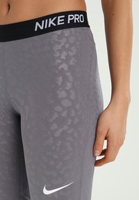 Szare legginsy Nike Pro mają delikatny wzór w panterkę, czarny pas w talii z napisem "NIKE PRO" oraz białe logo na nodze.