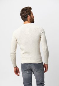 Cremfarbener Pullover mit langen Ärmeln und rundem Ausschnitt, der ein strukturiertes Strickmuster aufweist. Getragen mit grauen Jeans, von hinten betrachtet.