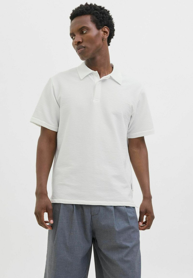 Hvid polo shirt med et tekstureret mønster, der har klassisk krave og korte ærmer, parret med løse grå bukser.