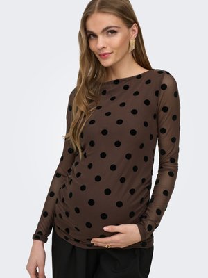 Femme enceinte en haut marron à manches longues avec des pois noirs, tenant doucement son ventre, portant des boucles d'oreilles et des bagues en or.