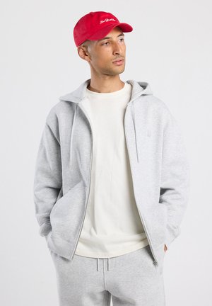 Sudadera con cremallera de color gris claro con bolsillos frontales, combinada con una camiseta blanca y pantalones de chándal grises. Gorra roja con texto blanco. Textura suave.
