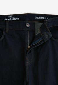 Dunkelblaue Denim-Jeans mit einem Reißverschluss, braunen Nähten und einem Knopfverschluss. Das Innere zeigt einen helleren Besatzstoff.