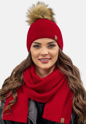 Beanie rosso lavorato a maglia con un pompon in pelliccia sintetica e una sciarpa coordinata con texture, entrambi caratterizzati da un design a costine e un piccolo etichetta di marca.
