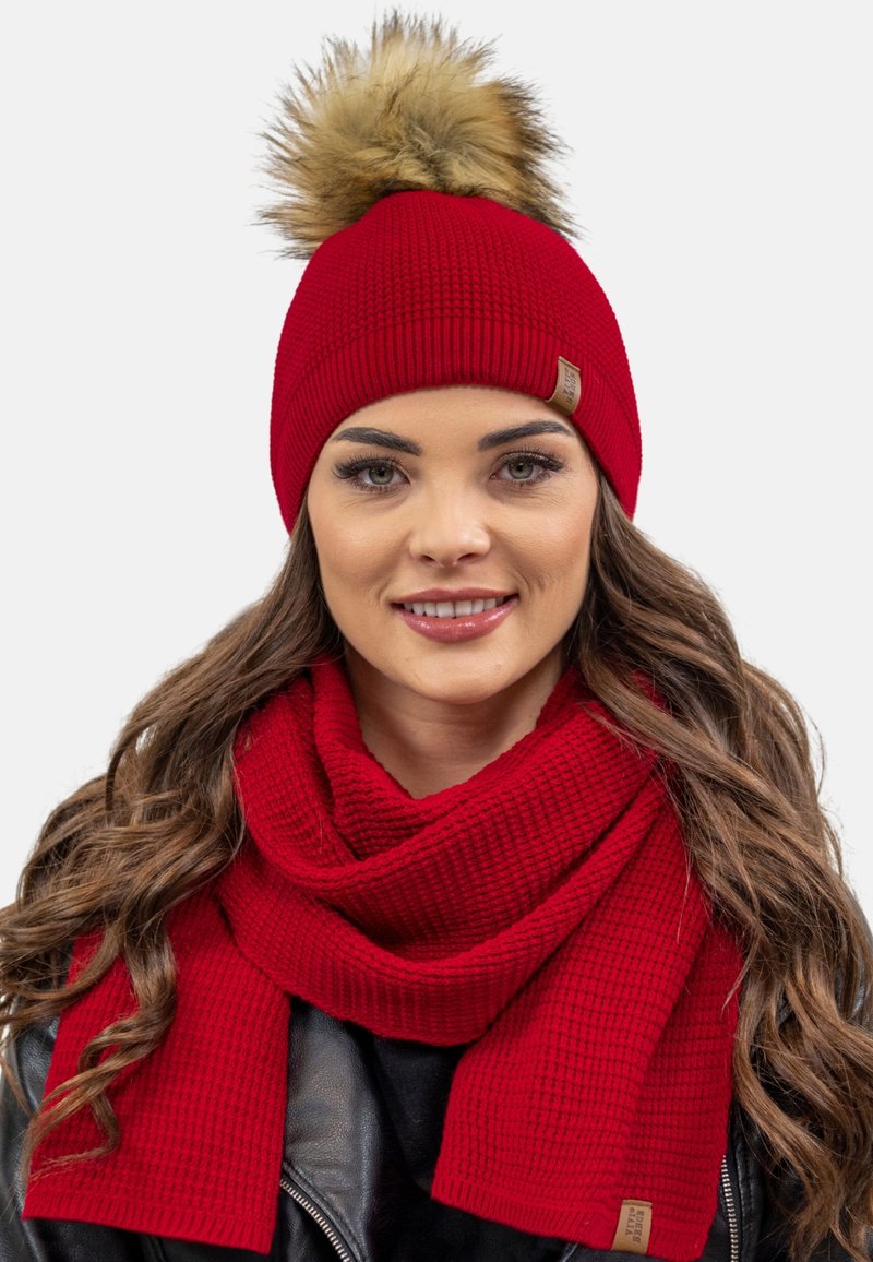 Căciulă tricotată roșie cu un pom-pom din blană sintetică și o eșarfă texturată asortată, ambele având un design cu nervuri și o etichetă mică cu marcă.