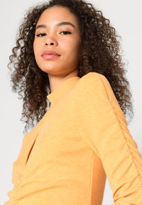 Top canelado de mangas longas em laranja brilhante, com um decote alto e detalhes franzidos nas laterais para um toque adicional de textura.