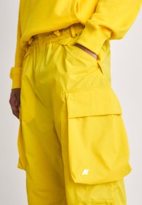 Pantaloni cargo gialli realizzati in materiale impermeabile, con grandi tasche laterali, cintura elastica e dettaglio riflettente del logo.