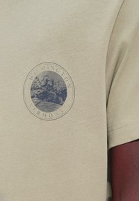 Κοντινό πλάνο μανικιού μπεζ πουκάμισου με κυκλικό γραφικό που δείχνει δέντρα και τοπίο, με την επιγραφή Wilmington Vermont στο στήθος.