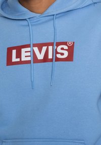 Ljusblå hoodie i mjukt tyg med en röd rektangulär logotyp som har "LEVI'S" i vita fetstilta bokstäver; inkluderar snörning.