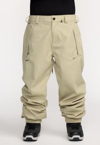 Beige sneeuwbroeken met een relaxed fit, voorzien van ritssluitingen in de zakken, een verstelbare tailleband en elastische manchetten, gecombineerd met zwarte laarzen.