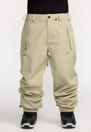 Pantalons de neige beiges à coupe décontractée, avec poches zippées, taille ajustable et poignets élastiques, assortis à des bottes noires.