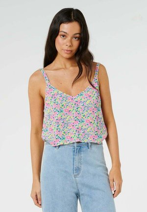 Vrouw met lang donker haar die een roze bloemenmouwloos topje en lichtblauwe jeans met hoge taille draagt, staand tegen een effen achtergrond.
