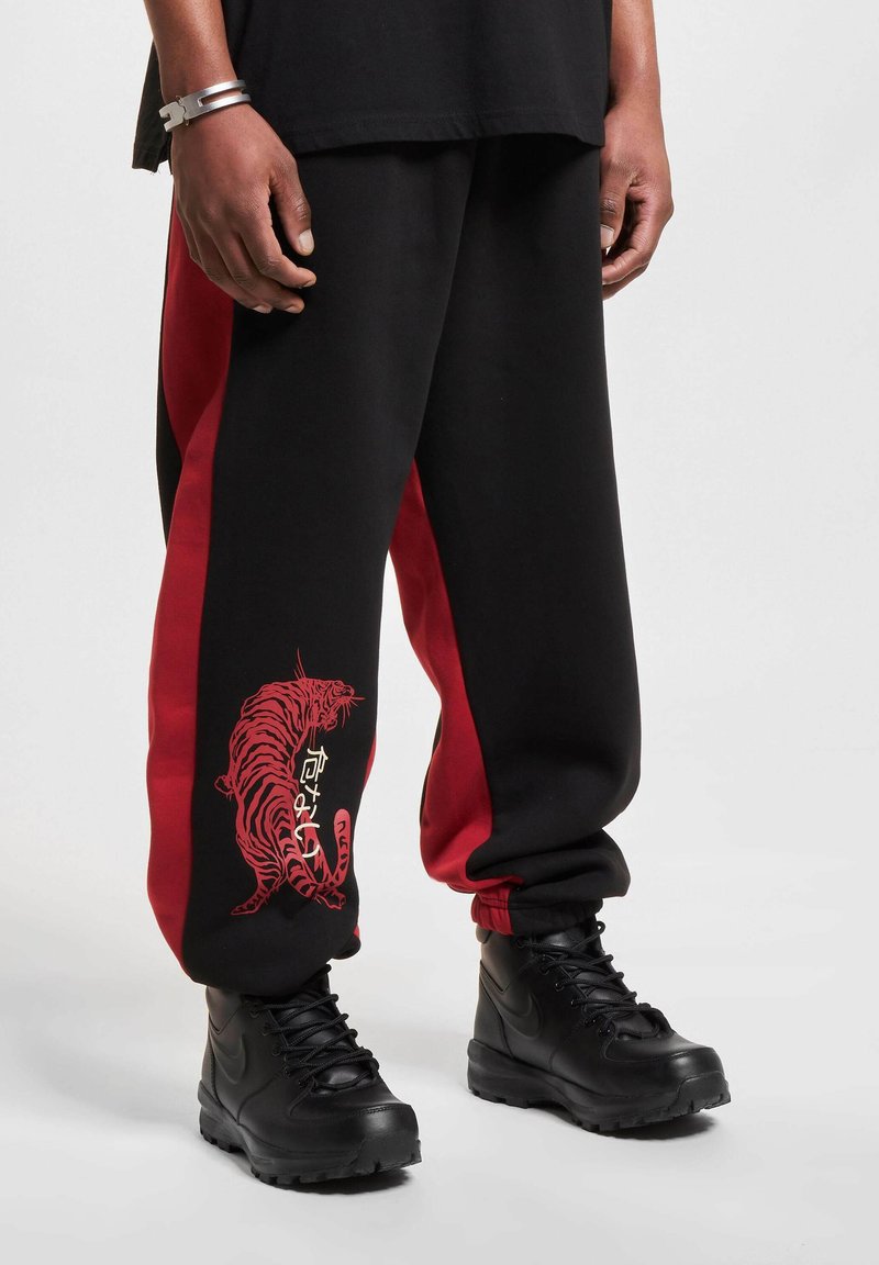 Black jogger pants with red side panels, featuring a red tiger graphic and text. Paired with black lace-up boots. Cotton material.