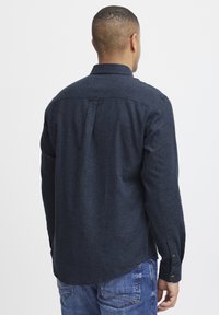 Camisa de manga larga azul marino hecha de tejido texturizado, con un cuello tipo polo y un pliegue en la parte central de la espalda, usada sobre unos vaqueros de mezclilla.