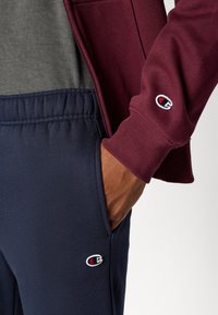 Bordeaux zip hoodie met een marineblauwe zak en marineblauwe sweatpants. Beide kledingstukken zijn gemaakt van zachte stof met geborduurde logo-accenten.