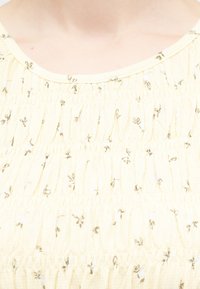 Κοντινό πλάνο υφάσματος ανοιχτού κίτρινου χρώματος με στρογγυλεμένες ραφές και μικρά καφέ λουλουδένια σχέδια σε ρούχο με στρογγυλή λαιμόκοψη.