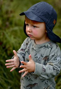 Blå solhatt i tyg med flikar; grå långärmad tröja med flerfärgade dinosauriemönster; barn med ljust brunt hår som gestikulerar med händerna.