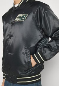 Personne portant une veste bomber en satin noir avec des poignets et un bas à rayures blanches, main dans la poche, et un logo "NB" vert et blanc sur la poitrine.