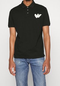 Svart pikétröja i bomull med en treknappsknäppning och korta ärmar. Har ett vitt hjärta och "V"-logotyp på vänster bröst.