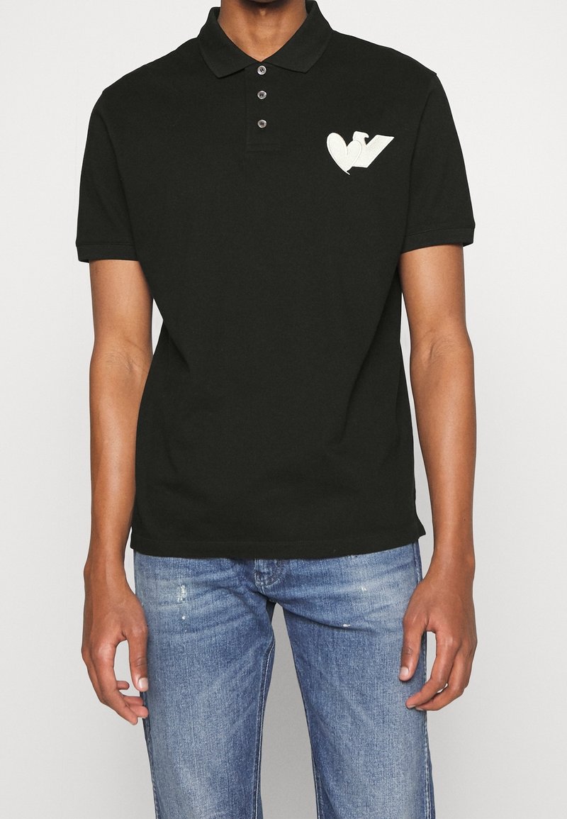 Svart pikétröja i bomull med en treknappsknäppning och korta ärmar. Har ett vitt hjärta och "V"-logotyp på vänster bröst.