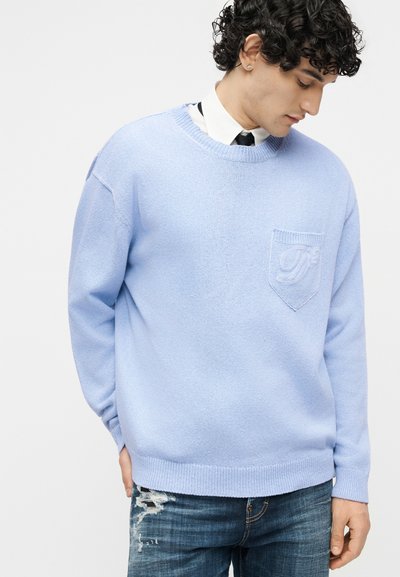 Jeune homme aux cheveux bouclés portant un pull bleu clair sur une chemise blanche avec une cravate noire, associé à un jean bleu déchiré.