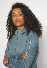 Woman with curly hair wearing a gray zip-up jacket with a blue zipper detail on the arm, arms crossed, looking forward.