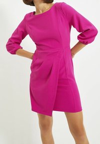 Vestido fucsia con mangas abullonadas de tres cuartos, cuerpo ajustado, detalles plisados en la cintura y corte recto por encima de las rodillas.