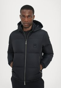 Black puffer jacket with a hood, featuring quilted sections, front zipper closure, and logo patch on the chest. Soft, water-resistant material.