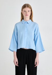 Camicetta azzurra chiara corta con superficie testurizzata, chiusura con bottoni e colletto. Presenta maniche larghe e una vestibilità rilassata, abbinata a pantaloni neri.