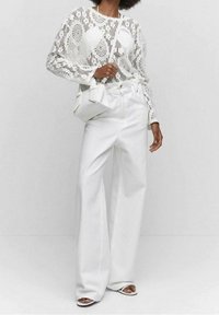 Bílý krajkový top s dlouhými rukávy a propracovanými vzory, kombinovaný s vysokým pasem a širokými nohavicemi bílých kalhot. Doplňkem je malá bílá kabelka.