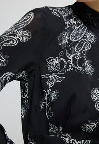 Zwarte blouse met witte bloemen- en paisleypatronen, gemaakt van lichte, gestructureerde stof; heeft een hoge kraag en lange mouwen.