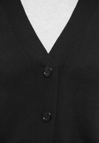 Svart cardigan med V-halsdesign, med to svarte knapper foran og ribbestruktur. Bakgrunnen viser en gråstripete skjorte.