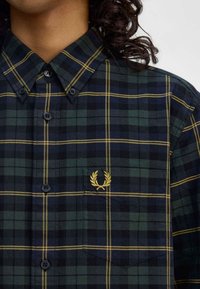Grün- und marineblau karierter Button-Up-Hemd mit einem gelben gestickten Logo, ausgestattet mit einem Kragen und einer Brusttasche mit einem spitzen Klappdeckel.