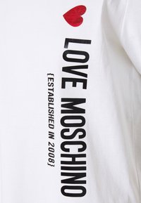 Vit T-shirt med en stor svart tryckt text "LOVE MOSCHINO" och en röd hjärtgrafik. Inkluderar mindre text "ETABLERAD 2008."