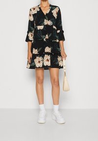 Vestido floral negro con grandes flores beiges y verdes, diseño en capas, escote en V, mangas 3/4, combinado con zapatillas blancas y un bolso crema.