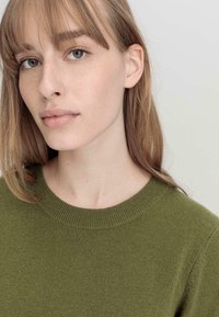 Maglione lavorato a maglia di colore verde scuro con scollo rotondo, texture morbida e bordi a costine, indossato da una persona con capelli castano chiaro e trucco naturale.