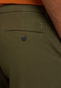 Oliva gröna byxor med en texturerad tyg, med en bakficka med en blå knapp och en broderad logotyp ovanför ficköppningen.