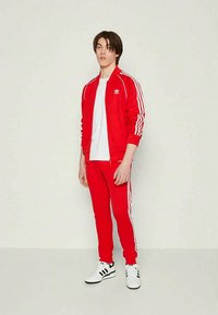 Jauns vīrietis stāv, ģērbies sarkanā Adidas sporta tērpā ar baltām svītrām un baltā T-kreklā, apavi - baltas Adidas kedas.