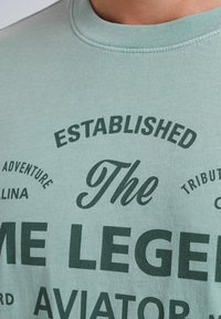 Světle zelené bavlněné tričko se tmavě zeleným textem, včetně "ZAŘÍZENÝ" a "LEGENDY," s různými styly písma. Kulatý výstřih.