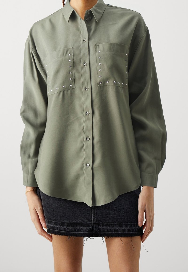 Chemise kaki à boutons avec manches longues, deux poches ornées de clous argentés, et un ourlet arrondi, associée à une jupe en denim noire.