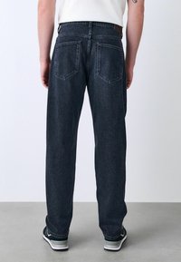Persona vestida con jeans rectos negros y zapatillas negras, de pie sobre un suelo gris contra un fondo liso, vista desde atrás.