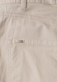 Gros plan sur un tissu beige avec une poche cousue et une petite étiquette métallique portant la mention "Street One" près de la couture de la poche.