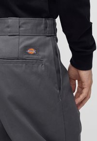 Záber na osobu nosiacu tmavosivé nohavice Dickies so zadným vreckom a logom, ruka spočívajúca pri bokoch, viditeľná čierna dlhá košeľa.