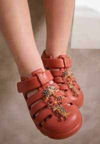 Orange rubber sandals featuring horizontal cutouts, adjustable strap, and three colorful dinosaur-shaped ornaments on the front.