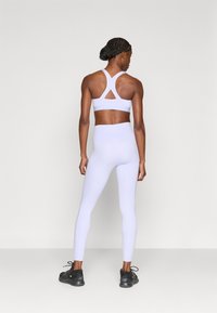 Αθλητικό σουτιέν σε ανοιχτό λεβάντα με τριγωνικό άνοιγμα στην πλάτη, συνδυασμένο με ψηλόμεσα κολάν σε ταιριαστό χρώμα και μαύρα αθλητικά παπούτσια.