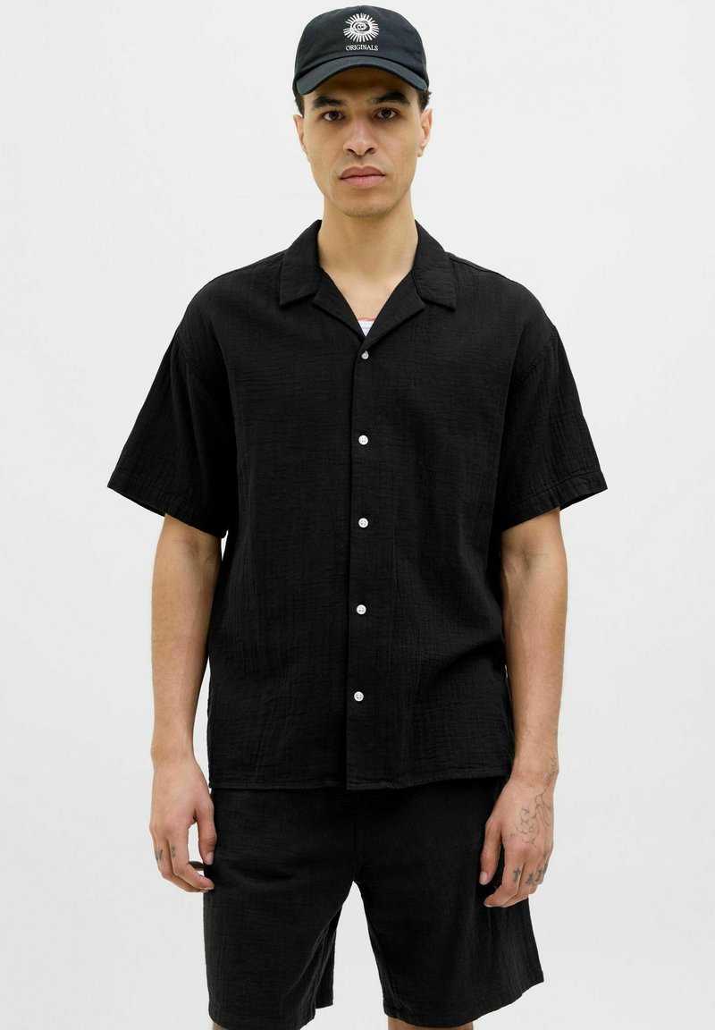 Camisa negra de manga corta con botones, hecha de un tejido texturizado, combinada con pantalones cortos a juego, con botones blancos y cuello.
