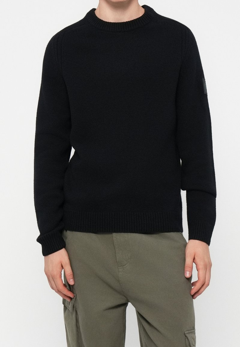 Personne portant un pull noir à col rond et un pantalon cargo vert olive, mains détendues le long du corps, sur un fond clair uni.
