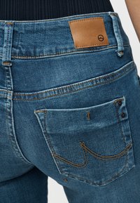 Κοντινό πλάνο πίσω τσέπης μπλε τζιν με διακοσμητική πορτοκαλί ραφή και καφέ δερμάτινο ταμπελάκι στη ζώνη μέσης.