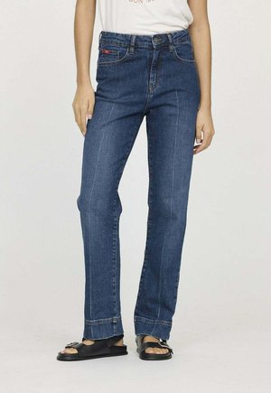 Femme portant un jean droit bleu et des sandales noires à bout ouvert, debout devant un fond uni.