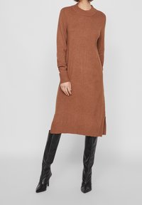 Vestito marrone lavorato a maglia con scollo rotondo, maniche lunghe, spacco laterale e dettagli a coste su polsini e orlo. Indossato con stivali neri al ginocchio.