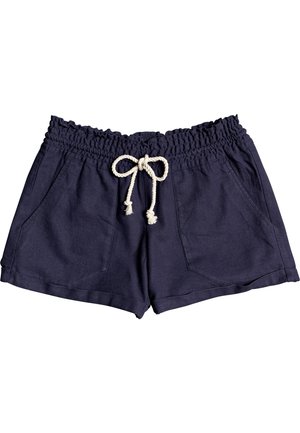 Kvinde iført hvid ærmeløs top med bladmotiv, marineblå shorts med snøre og lyse sneakers, stående mod en hvid baggrund.